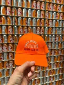 Een hand houdt een oranje Jopen Mooie Nel Pet (unisex) vast, met op de achtergrond een muur vol blikjes Jopen North Sea IPA-bier.