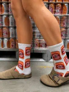 Een persoon draagt lichtbruine sandalen en een paar Jopen-sokken ‘Mooie Nel’: unisex-sokken – wit, met opvallende rode en oranje prints van sardineblikjes – en staat op beton voor stapels Open-sardineblikjes. Alleen de onderbenen zijn zichtbaar, waardoor dit unieke product perfect tot zijn recht komt voor fans van zowel sardines als opvallende sokken.