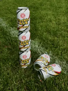 Een stapeltje van drie Tackle ALS-bierblikjes, met daarnaast nog twee blikjes die op het groene gras liggen.